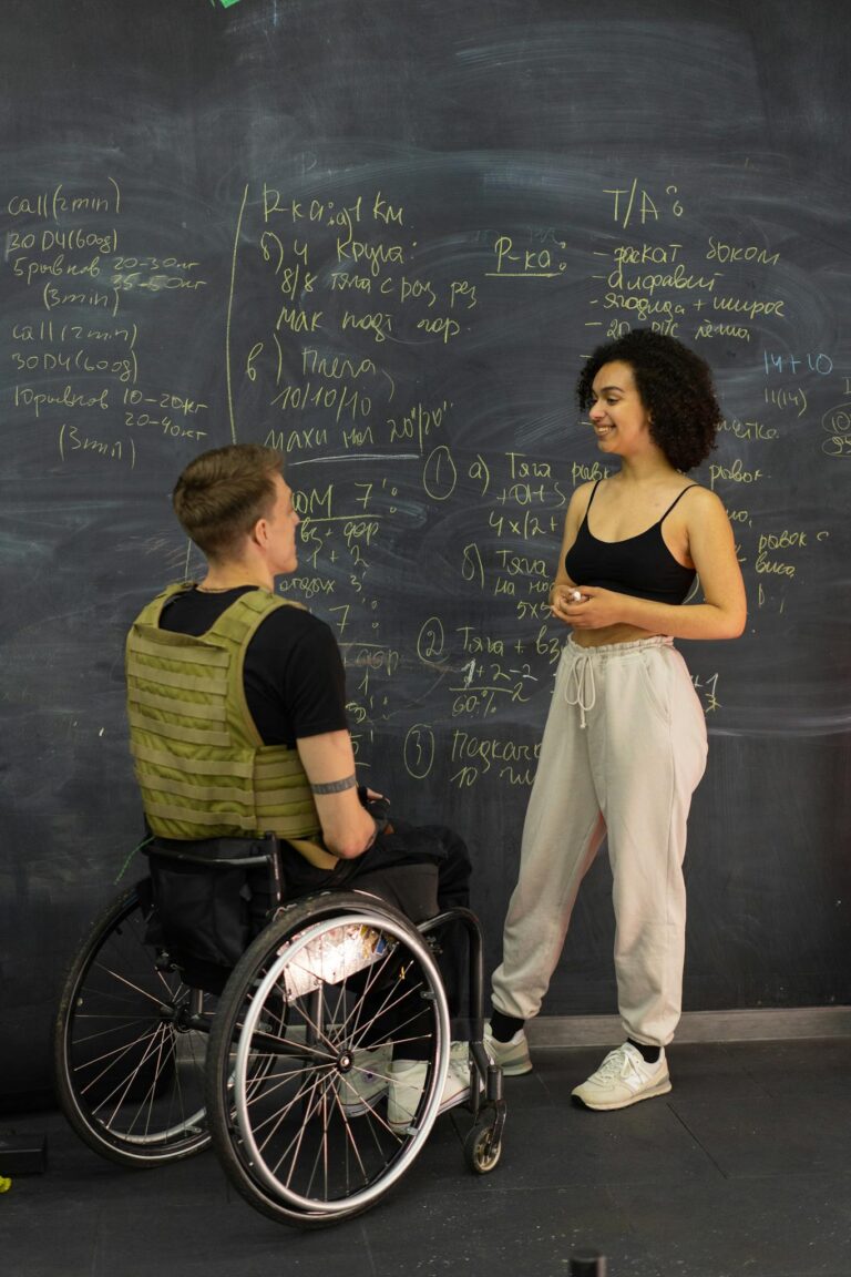 A female trainer and a disabled man in wheelchair discuss workout plans in gym setting.