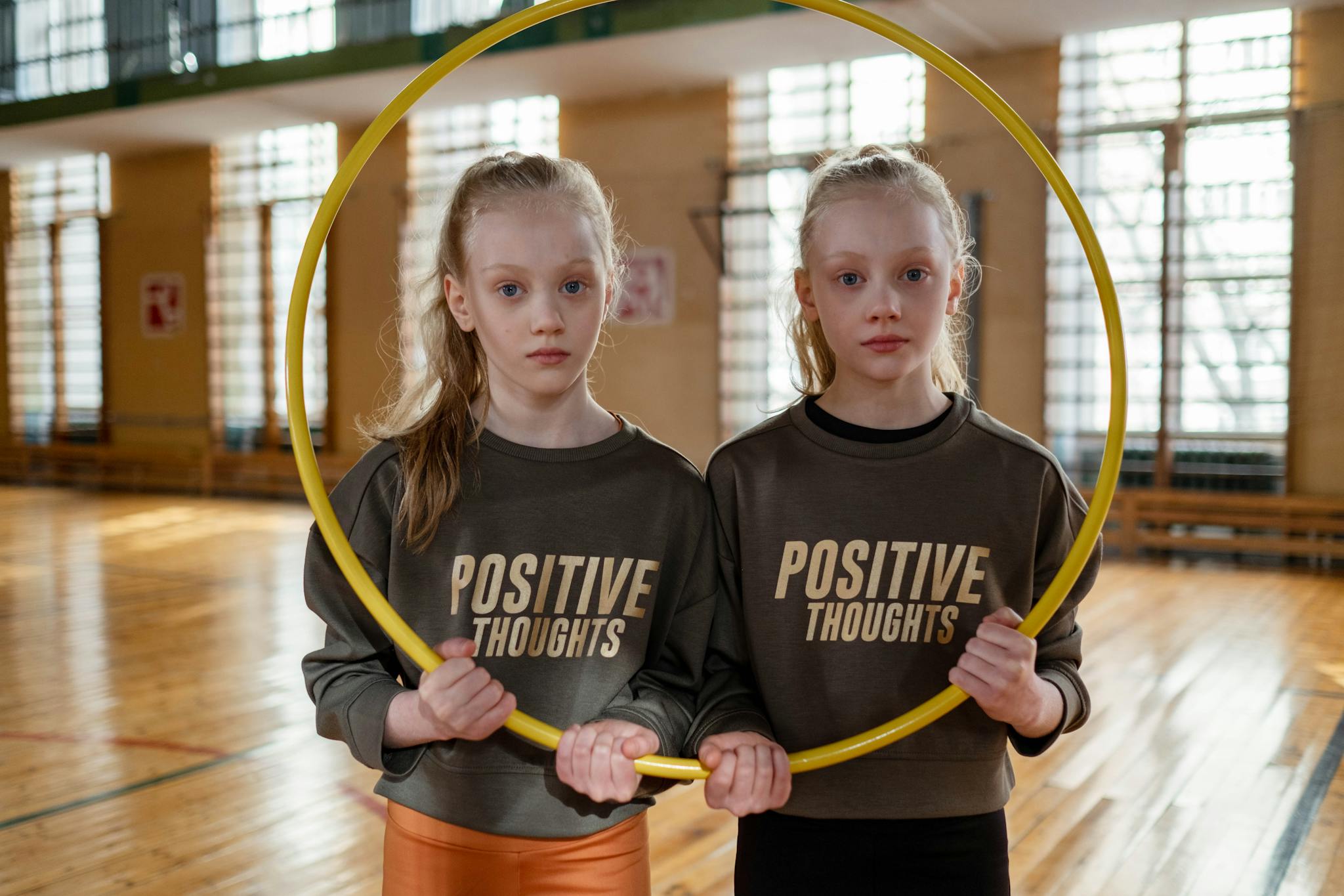Twin girls in gym holding a hula hoop, wearing 'Positive Thoughts' sweatshirts.